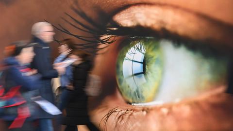 Messebesucher gehen an einer Wandtapete vorbei, auf dem ein menschliches Auge im Grossformat gedruckt ist. Messebesucher gehen an einer Wandtapete vorbei, auf dem ein menschliches Auge im Grossformat gedruckt ist.