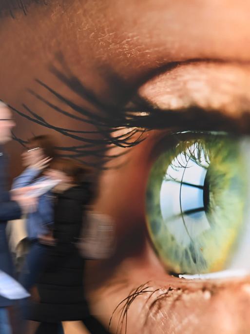 Messebesucher gehen an einer Wandtapete vorbei, auf dem ein menschliches Auge im Grossformat gedruckt ist.