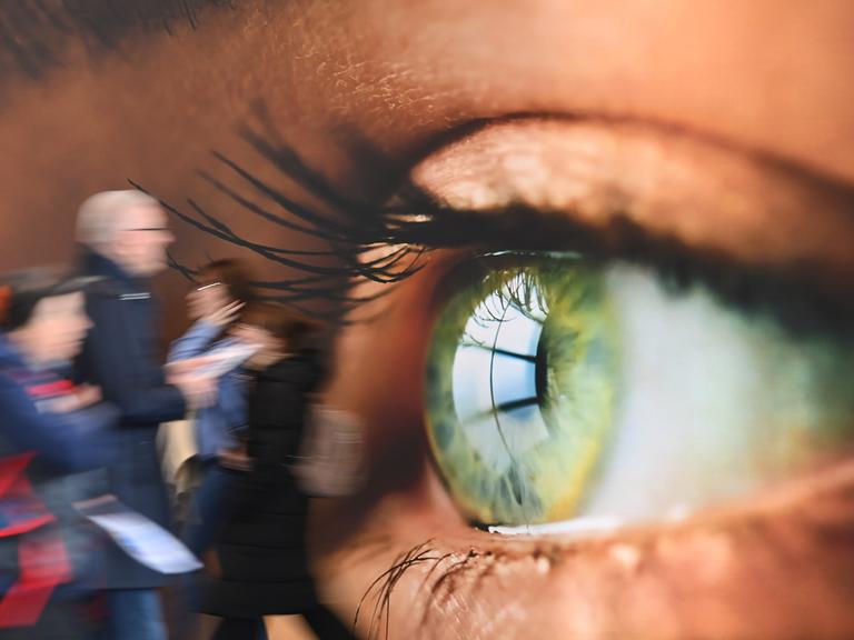 Messebesucher gehen an einer Wandtapete vorbei, auf dem ein menschliches Auge im Grossformat gedruckt ist.