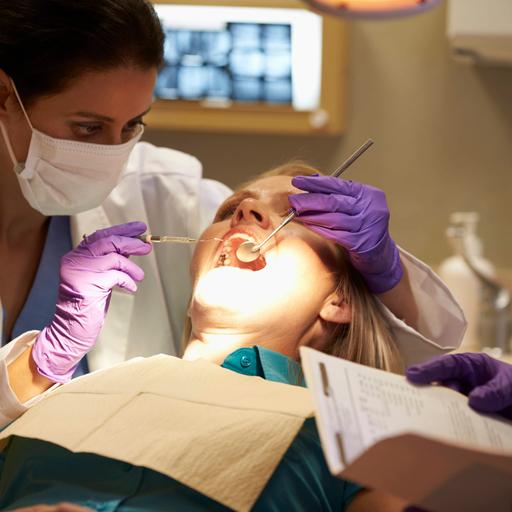 Eine Frau sitzt auf einem Behandlungsstuhl in einer Zahnarztpraxis. Sie hat den Mund offen und wird von einer Zahnärztin behandelt.