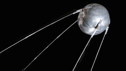 Abgebildet ist ein Modell des Satelliten Sputnik. Eine silberne Kugel, die von vier langen silbernen Streben gehalten wird.