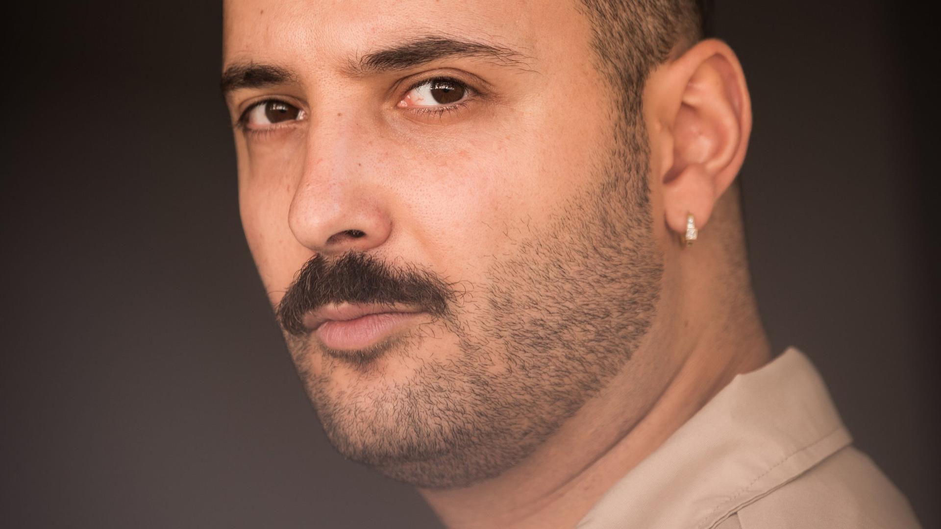 Fikri Anıl Altıntaş, ein junger Mann mit Schnurrbart und einem dezenten Ohrring im linken Ohr, schaut im Halbprofil entspannt in die Kamera.
