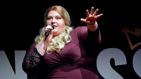 Nicole Jäger spricht auf der Bühne in ein Mikrofon. Sie hat blonde Locken und trägt ein Kleid in dunklem Lila.