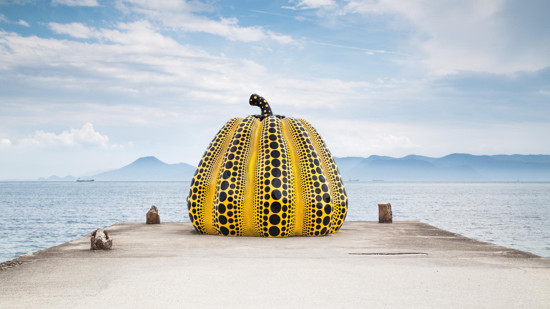 Auf einem Steg vor dem Meer steht eine mannshohe gelbe Kürbis-Skulptur, die mit schwarzen Punkten übersät ist.