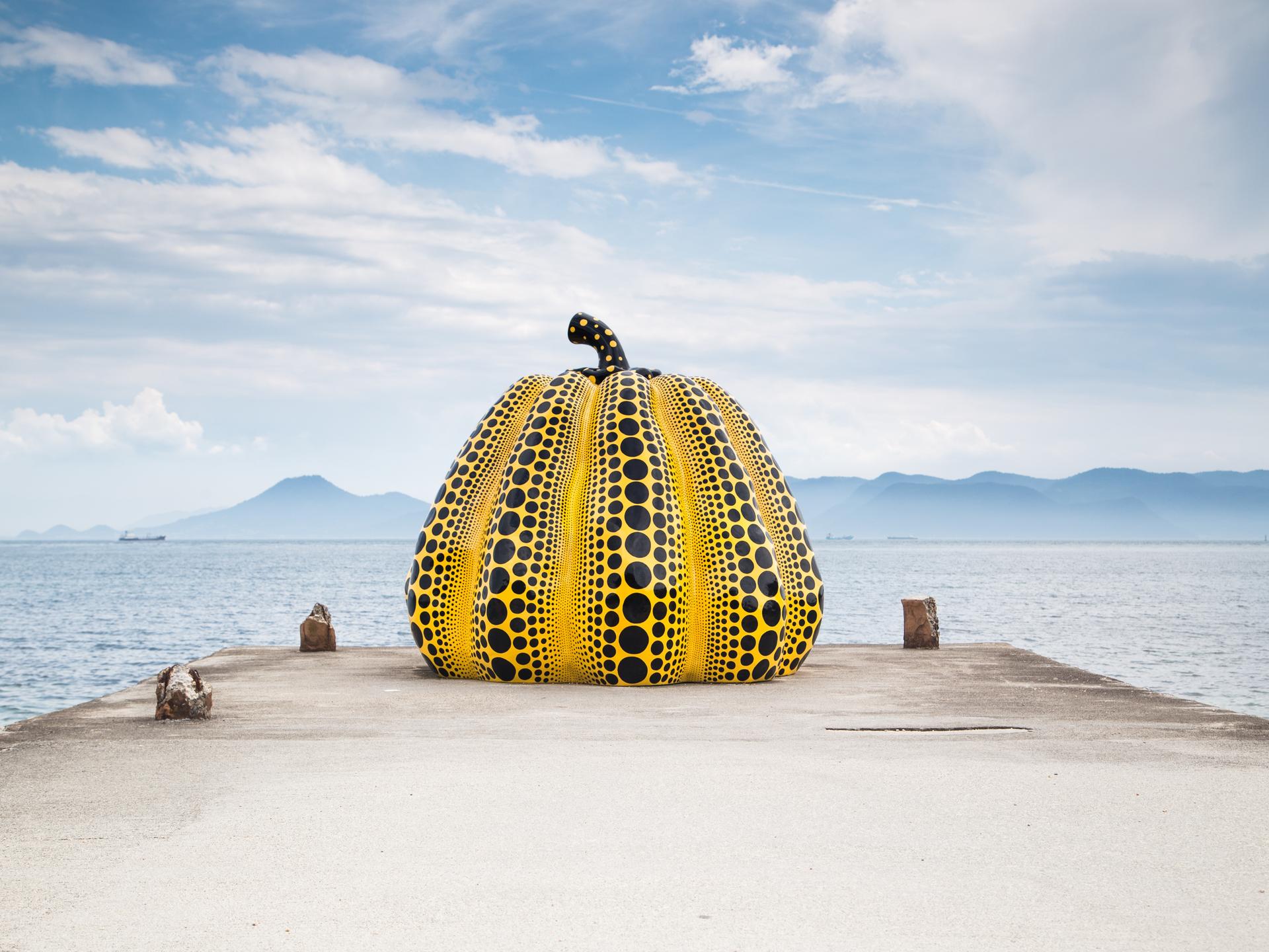 Auf einem Steg vor dem Meer steht eine mannshohe gelbe Kürbis-Skulptur, die mit schwarzen Punkten übersät ist. Auf einem Steg vor dem Meer steht eine mannshohe gelbe Kürbis-Skulptur, die mit schwarzen Punkten übersät ist.