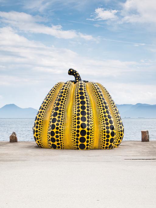 Auf einem Steg vor dem Meer steht eine mannshohe gelbe Kürbis-Skulptur, die mit schwarzen Punkten übersät ist. Auf einem Steg vor dem Meer steht eine mannshohe gelbe Kürbis-Skulptur, die mit schwarzen Punkten übersät ist.
