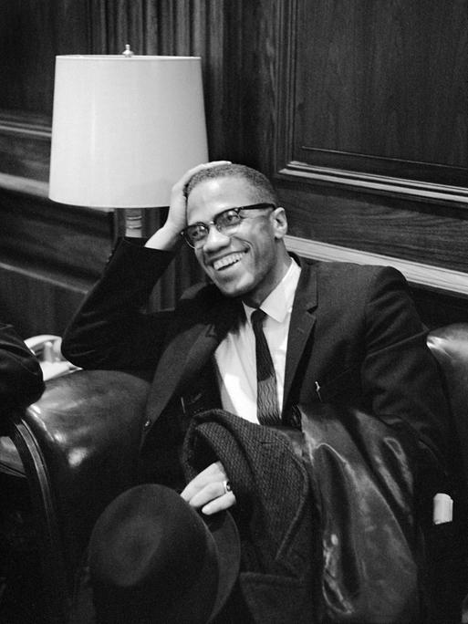 Der US-Bürgerrechtler Malcolm X im Anzug, er sitzt lächelnd auf einem Sessel und wartet auf die Pressekonferenz von Martin Luther King im Jahr 1964.