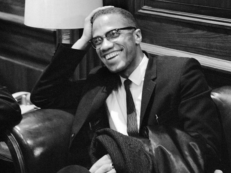 Der US-Bürgerrechtler Malcolm X im Anzug, er sitzt lächelnd auf einem Sessel und wartet auf die Pressekonferenz von Martin Luther King im Jahr 1964.
