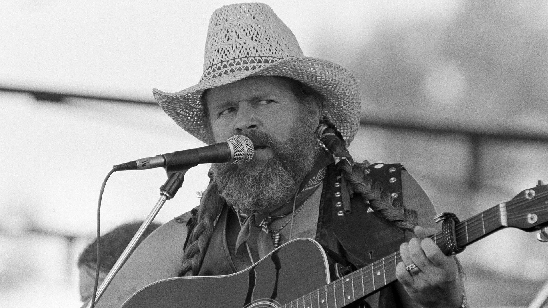 Der Country-Sänger David Allan Coe singt auf einem schwarz-weiß Foto mit Strohut und Gitarre in ein Mikrofon. 