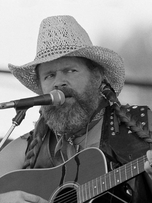 Der Country-Sänger David Allan Coe singt auf einem schwarz-weiß Foto mit Strohut und Gitarre in ein Mikrofon. 