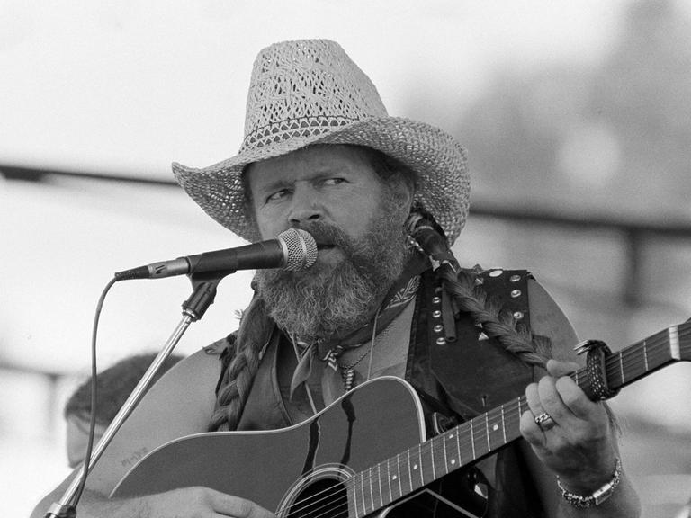 Der Country-Sänger David Allan Coe singt auf einem schwarz-weiß Foto mit Strohut und Gitarre in ein Mikrofon. 