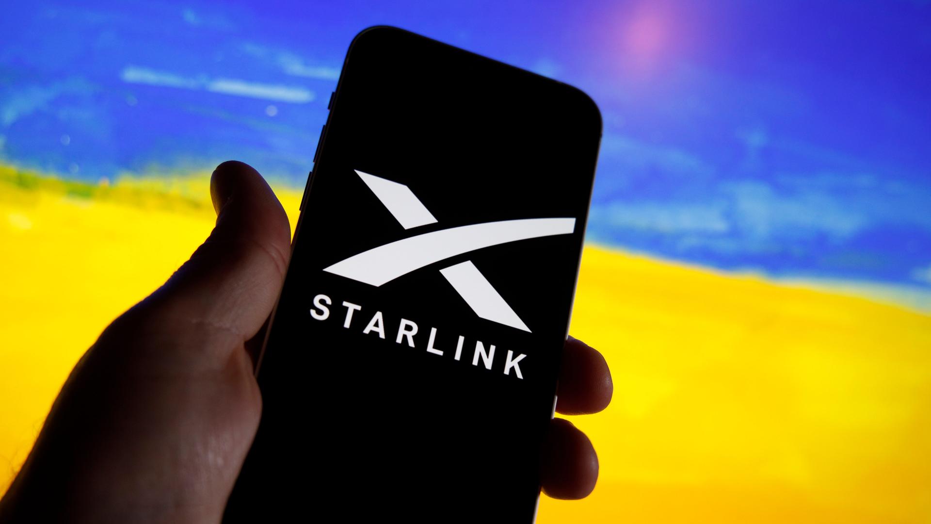 Ein Handy zeigt das Logo von Starlink. Im Hintergrund ist unscharf ein Feld mit gelben Blüten und ein blauer Himmel zu sehen, wodurch der Eindruck der ukrainischen Flagge erweckt wird.