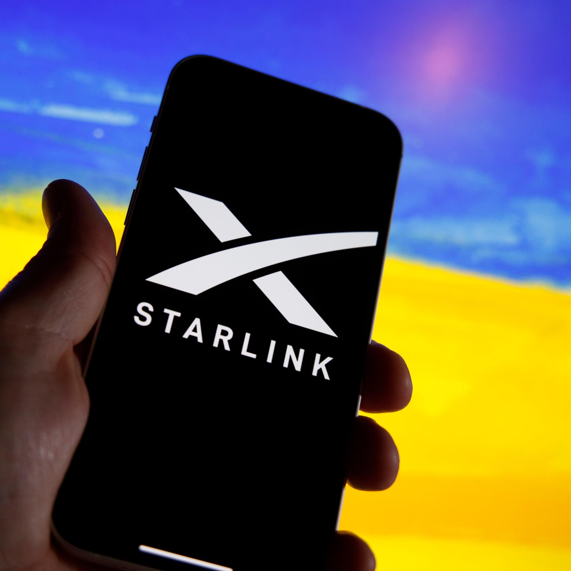 Ein Handy zeigt das Logo von Starlink. Im Hintergrund ist unscharf ein Feld mit gelben Blüten und ein blauer Himmel zu sehen, wodurch der Eindruck der ukrainischen Flagge erweckt wird.