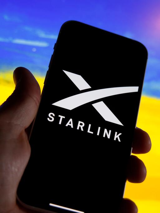 Ein Handy zeigt das Logo von Starlink. Im Hintergrund ist unscharf ein Feld mit gelben Blüten und ein blauer Himmel zu sehen, wodurch der Eindruck der ukrainischen Flagge erweckt wird.