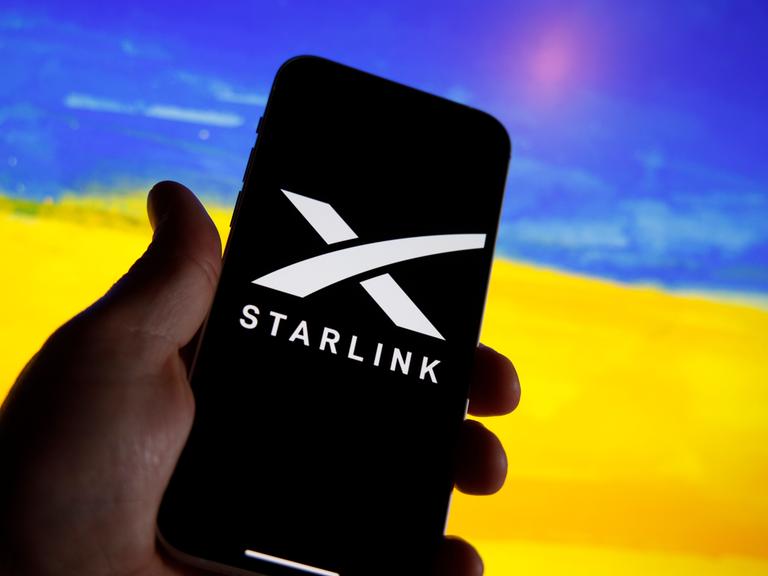 Ein Handy zeigt das Logo von Starlink. Im Hintergrund ist unscharf ein Feld mit gelben Blüten und ein blauer Himmel zu sehen, wodurch der Eindruck der ukrainischen Flagge erweckt wird.