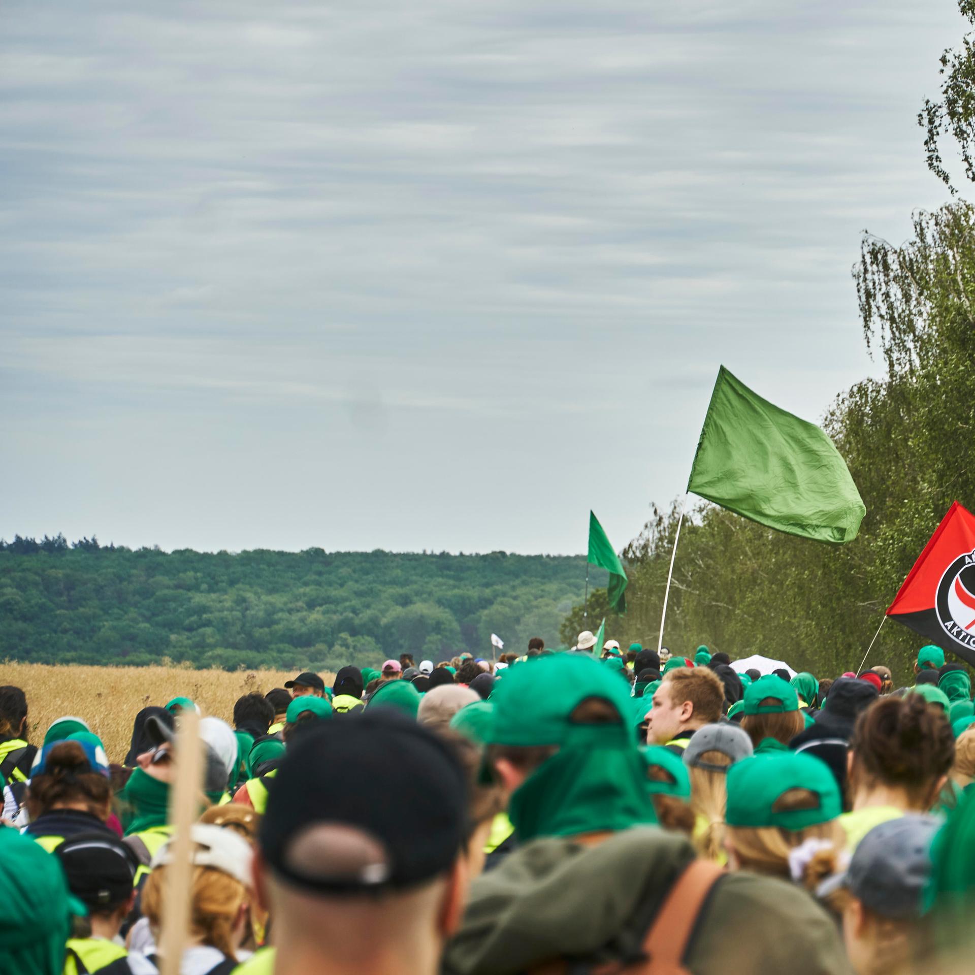Eine große Gruppe von Menschen laufen gemeinsam über Land. Die meisten tragen grüne Basecaps. Eine grüne Fahne wird geschwenkt, außerdem die Flagge der "Antifaschistischen Aktion".