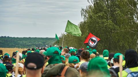 Eine große Gruppe von Menschen läuft gemeinsam über Land. Die meisten tragen grüne Basecaps. Eine grüne Fahne wird geschwenkt, außerdem die Flagge der "Antifaschistischen Aktion".