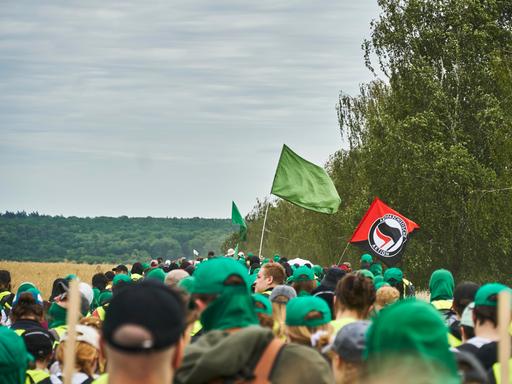Eine große Gruppe von Menschen läuft gemeinsam über Land. Die meisten tragen grüne Basecaps. Eine grüne Fahne wird geschwenkt, außerdem die Flagge der "Antifaschistischen Aktion". Eine große Gruppe von Menschen läuft gemeinsam über Land. Die meisten tragen grüne Basecaps. Eine grüne Fahne wird geschwenkt, außerdem die Flagge der "Antifaschistischen Aktion".