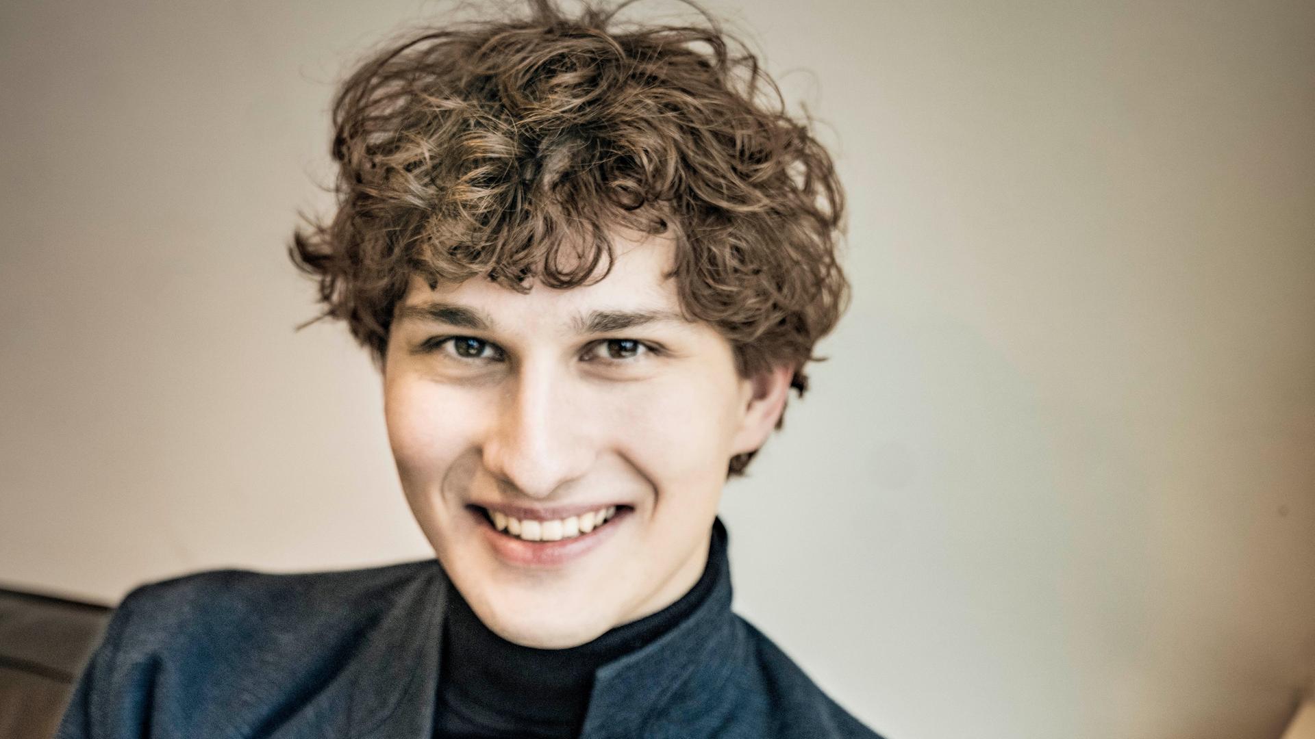 Freundlich lächelt ein junger Mann mit dunkelblondem Lockenkopf in die Kamera. Er sitzt auf einem Sofa und trägt einen schwarzen Rollkragenpullover und ein graues Wolljackett darüber.