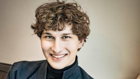 Freundlich lächelt ein junger Mann mit dunkelblondem Lockenkopf in die Kamera. Er sitzt auf einem Sofa und trägt einen schwarzen Rollkragenpullover und ein graues Wolljackett darüber.