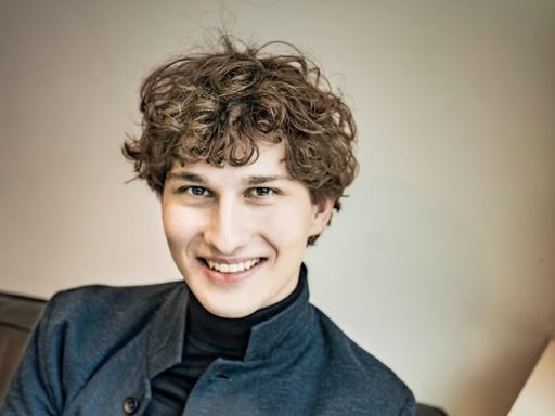 Freundlich lächelt ein junger Mann mit dunkelblondem Lockenkopf in die Kamera. Er sitzt auf einem Sofa und trägt einen schwarzen Rollkragenpullover und ein graues Wolljackett darüber.