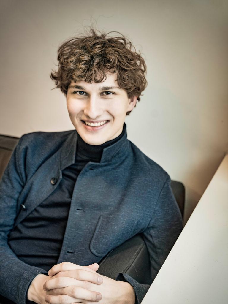Freundlich lächelt ein junger Mann mit dunkelblondem Lockenkopf in die Kamera. Er sitzt auf einem Sofa und trägt einen schwarzen Rollkragenpullover und ein graues Wolljackett darüber.