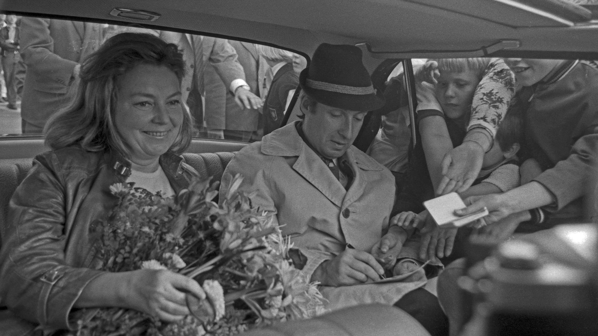 Schwarzweißbild von Peter Alexander und Hilde Haagen, die nebeneinander im Auto sitzen. Durch das Fenster stecken Fans Zettel mit Autogrammwünschen hinein.