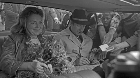Schwarzweißbild von Peter Alexander und Hilde Haagen, die nebeneinander im Auto sitzen. Durch das Fenster stecken Fans Zettel mit Autogrammwünschen hinein.
