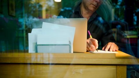 Ein Mann sitzt zu Hause an einem Schreibtisch. Er schreibt mit einem Stift auf ein Blatt Papier neben einem Laptop.