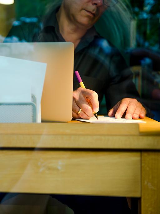 Ein Mann sitzt zu Hause an einem Schreibtisch. Er schreibt mit einem Stift auf ein Blatt Papier neben einem Laptop.