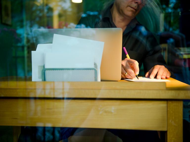 Ein Mann sitzt zu Hause an einem Schreibtisch. Er schreibt mit einem Stift auf ein Blatt Papier neben einem Laptop.