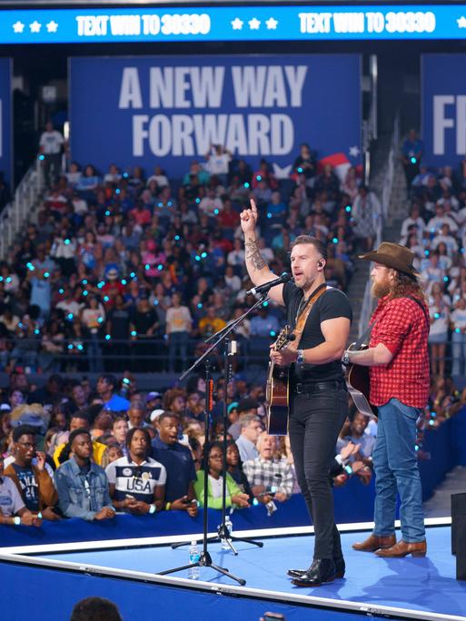 Ein Musiker mit einer Gitarre um den Hals und ein weiterer im roten Karohemd und mit Cowboy-Hut stehen auf einer Bühne vor einem großen Publikum. Im Hintergrund sind Banner mit den Worten "Freedom" und "A new way forward" zu sehen.