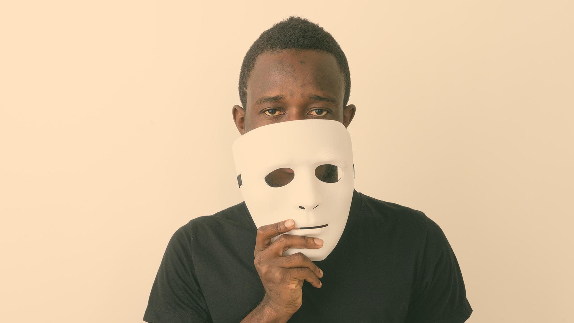 Ein schwarzer Mann hält eine weiße Maske vor das Gesicht. 