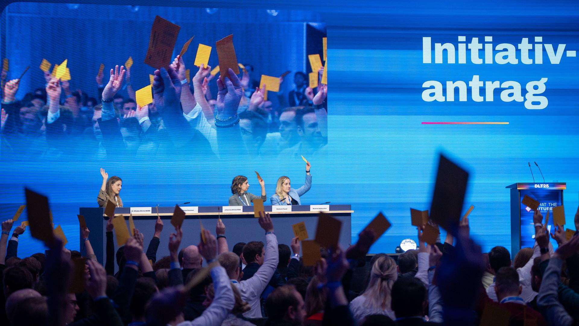 In einer Halle sitzen auf einem Podium auf dem Deutschlandtag der Jungen Union drei Personen, die die Hand gehoben haben oder Karten in die Höhe halten. Im Vordergrund sieht man Menschen von hinten im Publikum, die gelbe Karten in die Höhe halten. In einer Halle sitzen auf einem Podium auf dem Deutschlandtag der Jungen Union drei Personen, die die Hand gehoben haben oder Karten in die Höhe halten. Im Vordergrund sieht man Menschen von hinten im Publikum, die gelbe Karten in die Höhe halten.