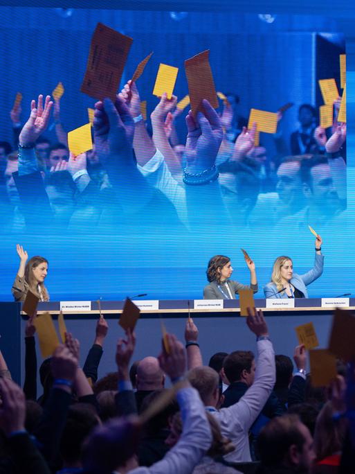 In einer Halle sitzen auf einem Podium auf dem Deutschlandtag der Jungen Union drei Personen, die die Hand gehoben haben oder Karten in die Höhe halten. Im Vordergrund sieht man Menschen von hinten im Publikum, die gelbe Karten in die Höhe halten.