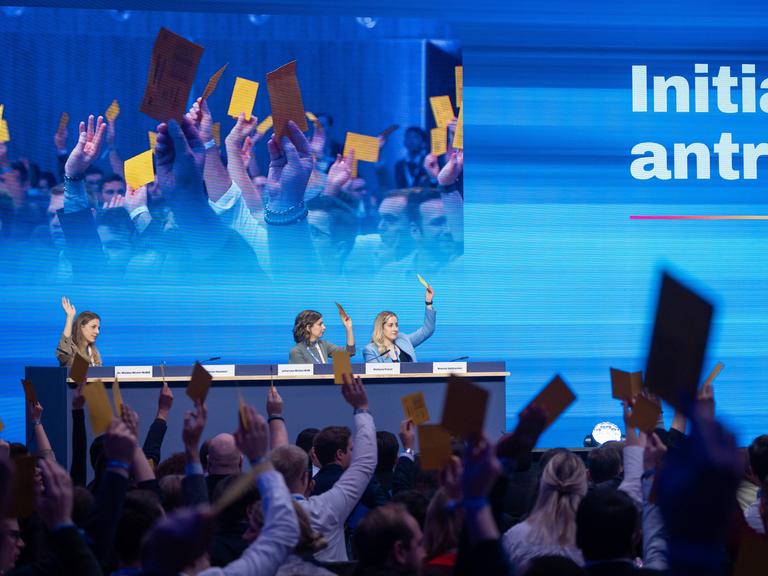 In einer Halle sitzen auf einem Podium auf dem Deutschlandtag der Jungen Union drei Personen, die die Hand gehoben haben oder Karten in die Höhe halten. Im Vordergrund sieht man Menschen von hinten im Publikum, die gelbe Karten in die Höhe halten.