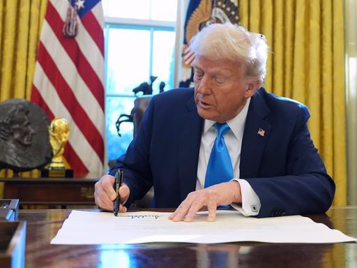 Donald Trump sitzt an einem Tisch im Weißen Haus und unterzeichnet ein Dokument.