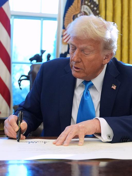 Donald Trump sitzt an einem Tisch im Weißen Haus und unterzeichnet ein Dokument.