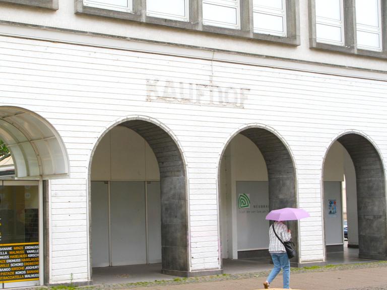 Zu sehen ist die geschlossene Filiale des Karstadt Kaufhof-Konzers in Neubrandenburg mitten in der Fußgängerzone. Davor läuft eine Passantin unter einem pinken Regenschirm.