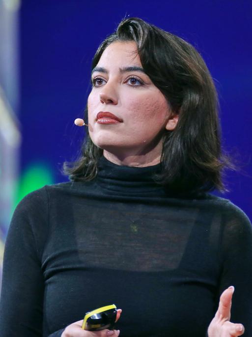 Eine Frau mit schwarzen Haaren steht auf der Bühne und schaut in das Publikum.