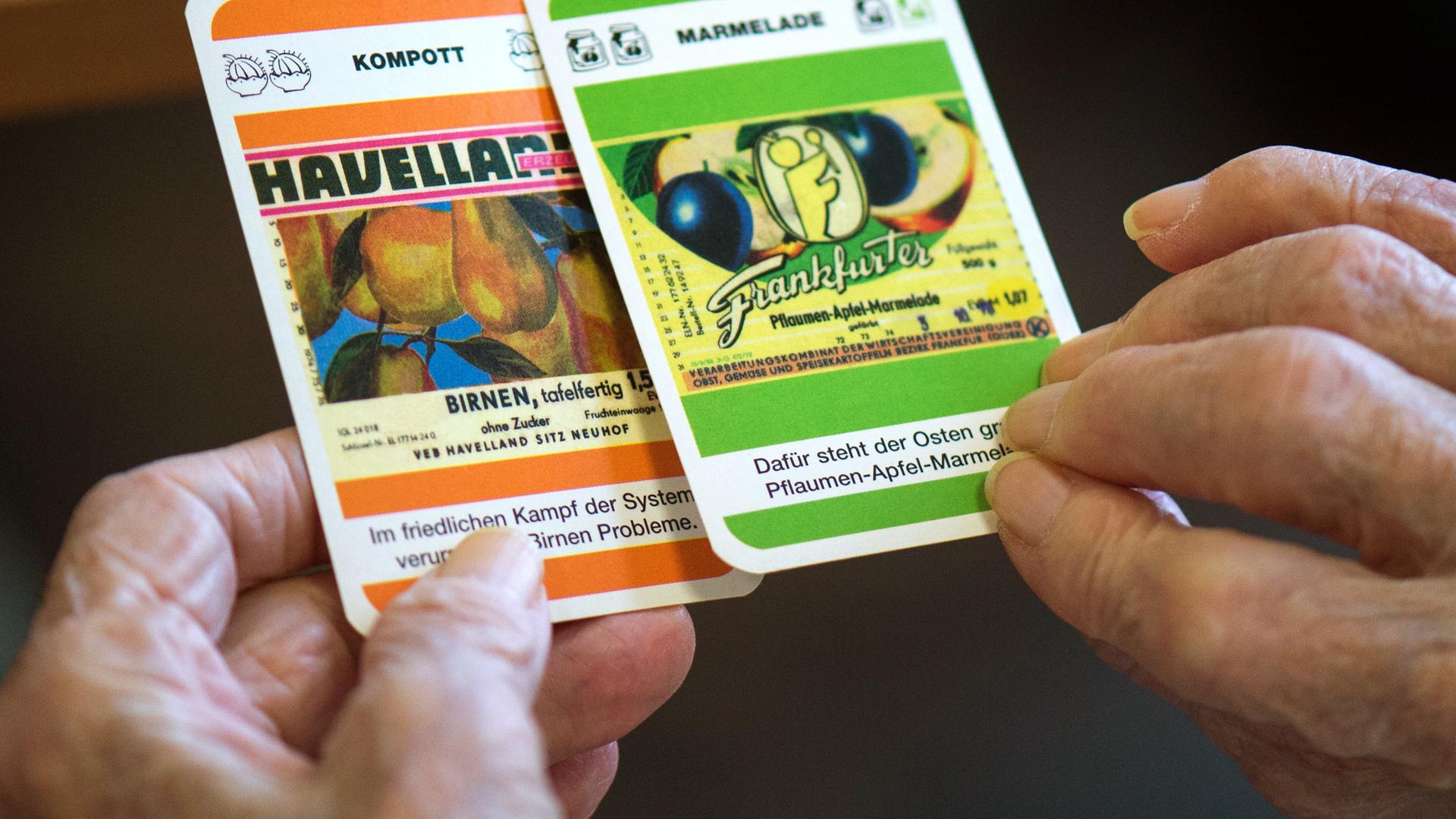 Zwei faltige Hände halten Spielkarten mit Fragen zu Produkten der DDR ("Kompott: Havelland").