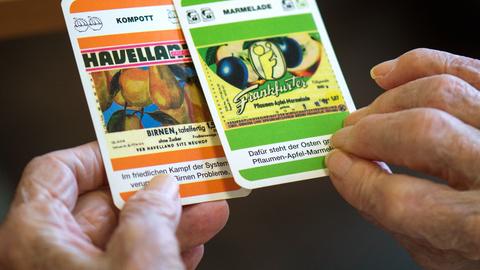 Zwei faltige Hände halten Spielkarten mit Fragen zu Produkten der DDR ("Kompott: Havelland"). Zwei faltige Hände halten Spielkarten mit Fragen zu Produkten der DDR ("Kompott: Havelland").
