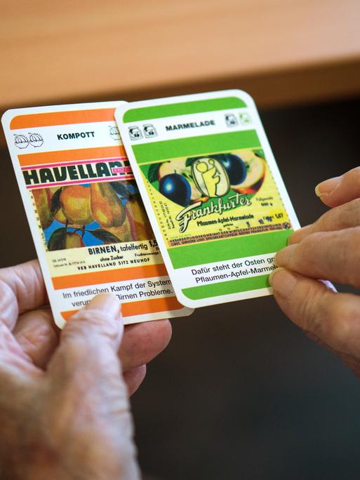 Zwei faltige Hände halten Spielkarten mit Fragen zu Produkten der DDR ("Kompott: Havelland").
