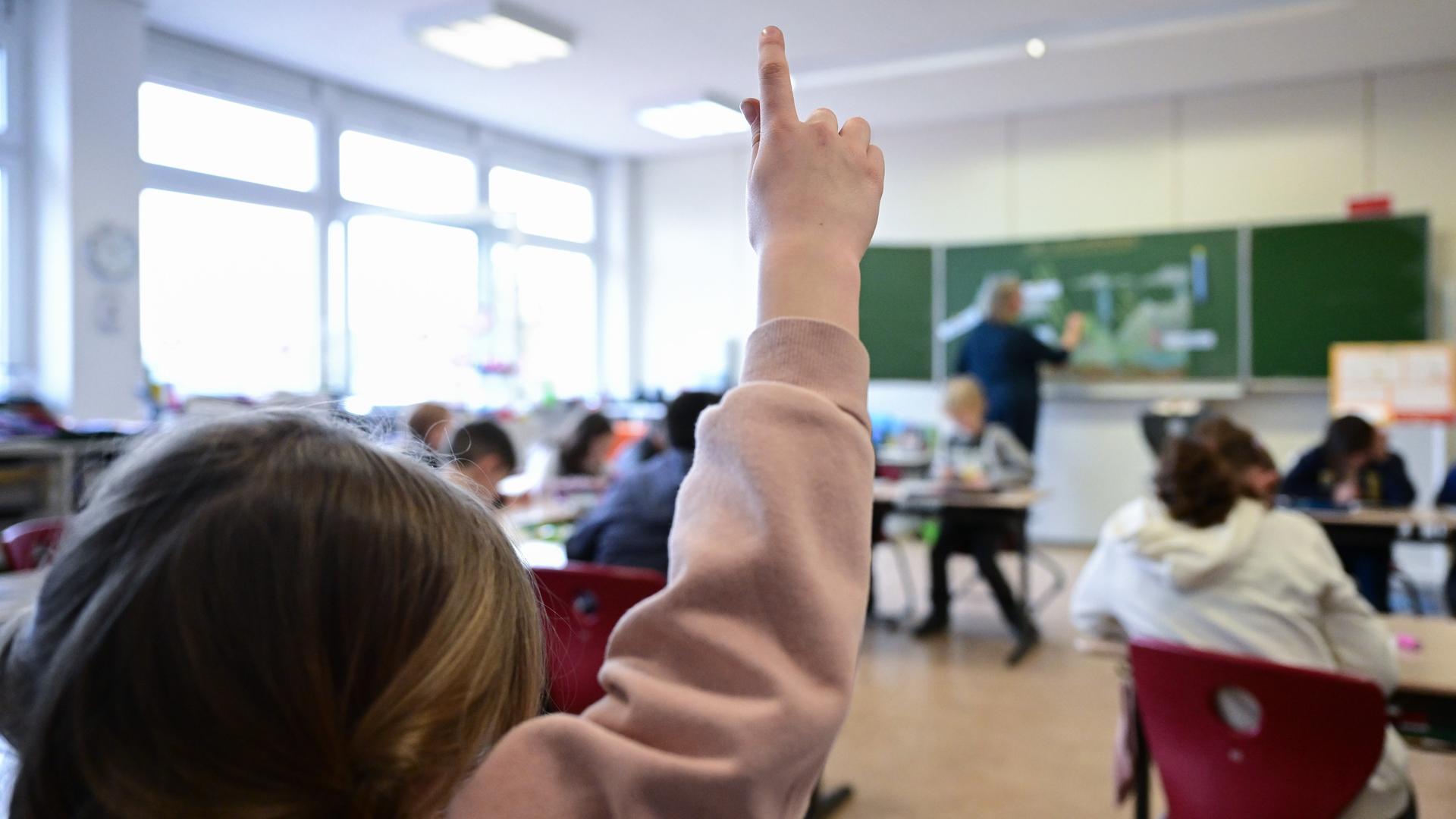 Schulkinder nehmen am Unterricht in einer Grundschule in Stuttgart teil. Ein Kind in rosafarbenem Pullover zeigt auf. Im Hintergrund des Bildes sieht man einen Lehrer an der Tafel stehen, der etwas mit Kreide notiert.