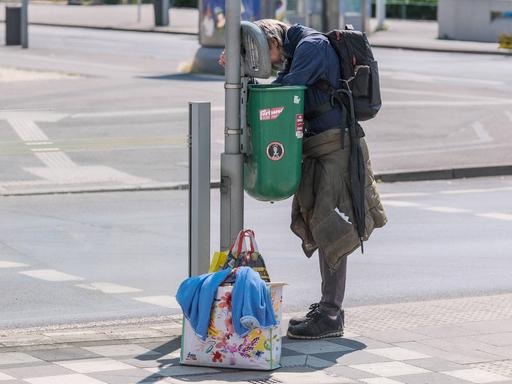 Ein Obdachloser guckt in einen Mülleimer, neben ihm stehen Tüten mit seinem Hab und Gut.