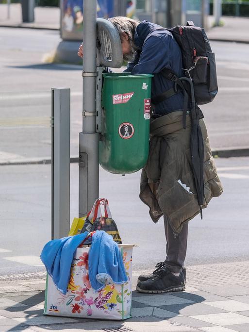 Ein Obdachloser guckt in einen Mülleimer, neben ihm stehen Tüten mit seinem Hab und Gut.