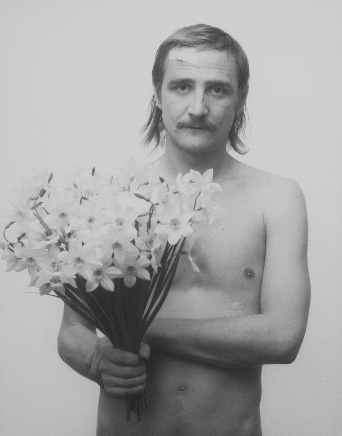 Das Schwarzweißfoto von Rudolf Bonvie zeigt einen Mann mit nacktem Oberkörper, der einen Strauß Narzissen in den Händen hält.