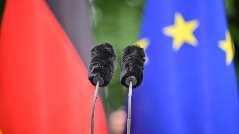 Zwei Mikrofone ragen aus einem Rednerpult. Im Hintergrund sind die deutsche und die EU-Flagge zu sehen. Zwei Mikrofone ragen aus einem Rednerpult. Im Hintergrund sind die deutsche und die EU-Flagge zu sehen.