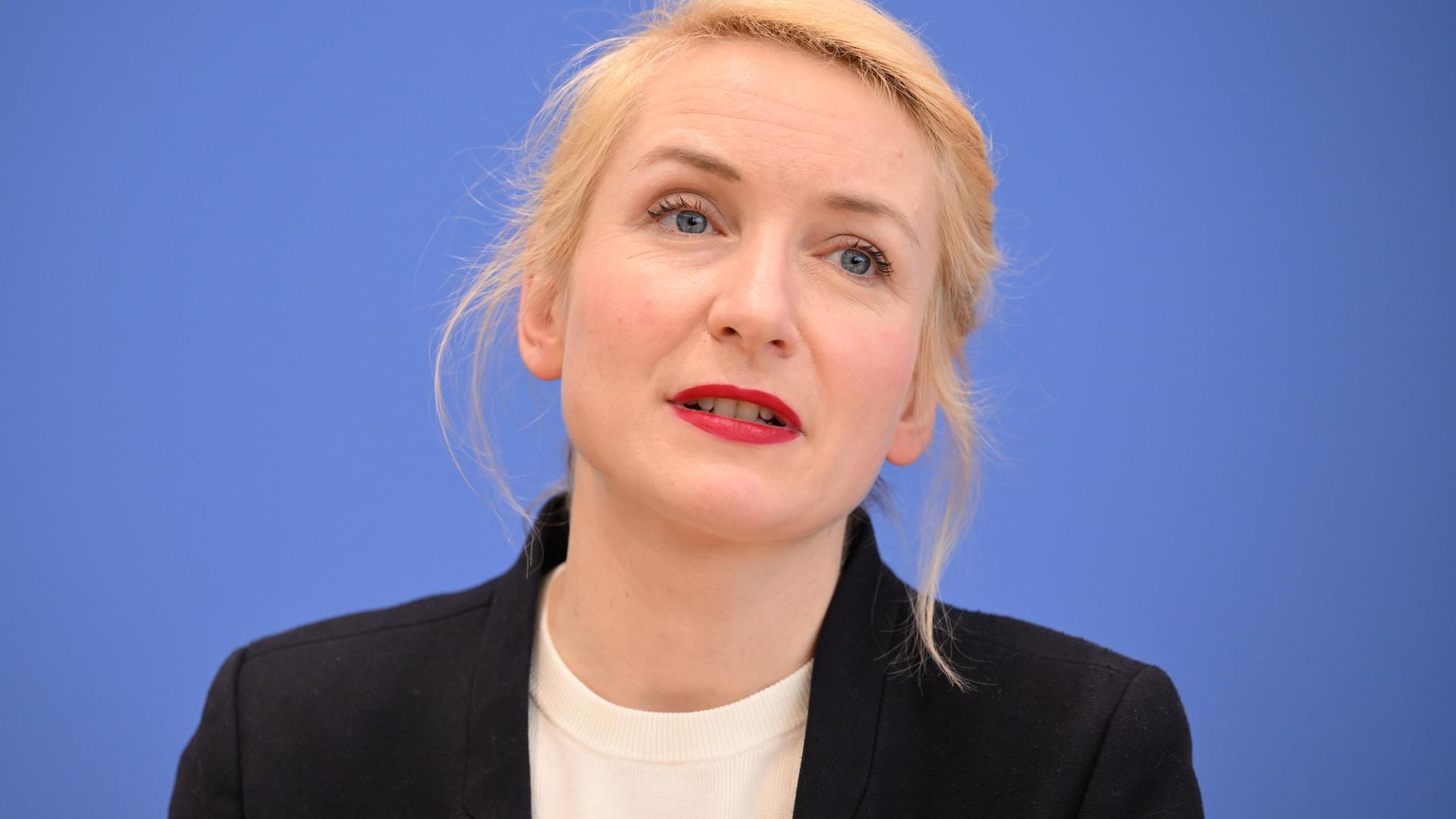 Ines Schwerdtner bei einer Pressekonferenz. Sie sitzt vor einem blauen Hintergrund und hat einen schwarzen Blazer an. 