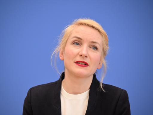 Ines Schwerdtner bei einer Pressekonferenz. Sie sitzt vor einem blauen Hintergrund und hat einen schwarzen Blazer an. 